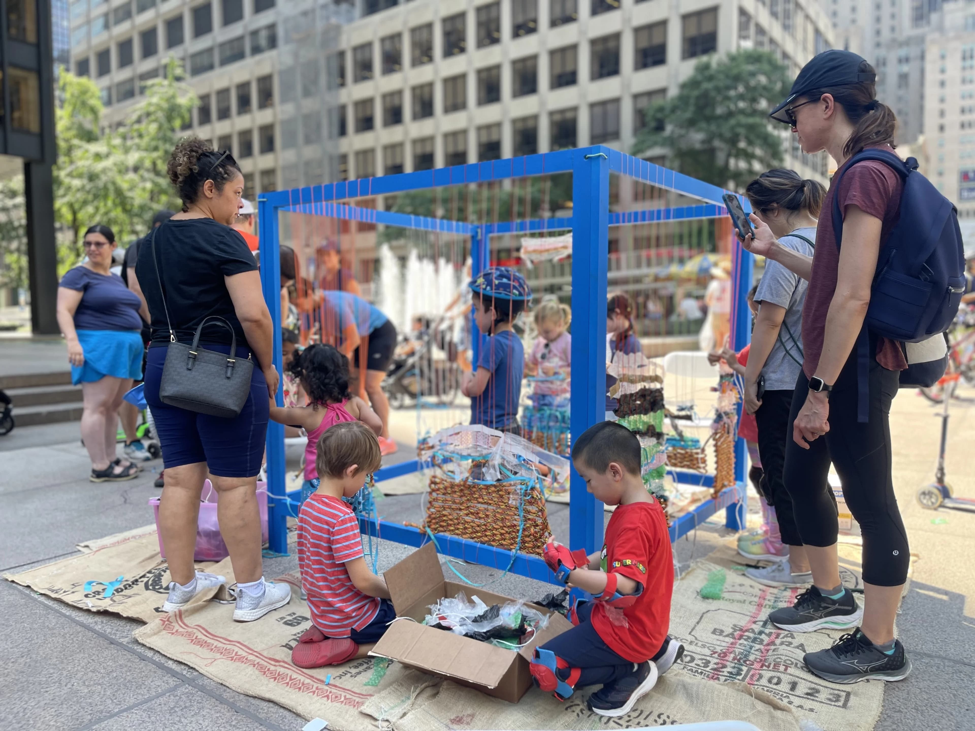 Children and families gathered around a blue collaborative loom during a NYC street weaving activation